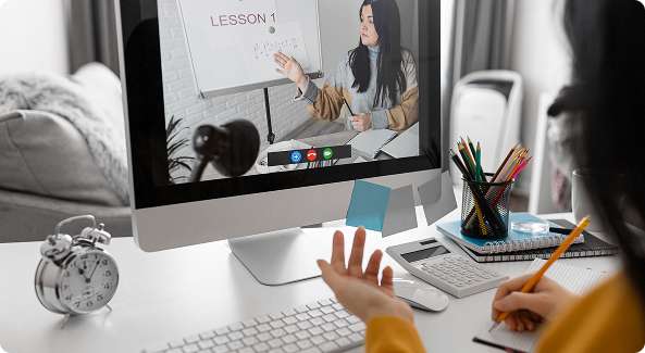 Student attending an online class on the best e-learning platform, taking notes at a desk with a computer and stationery.