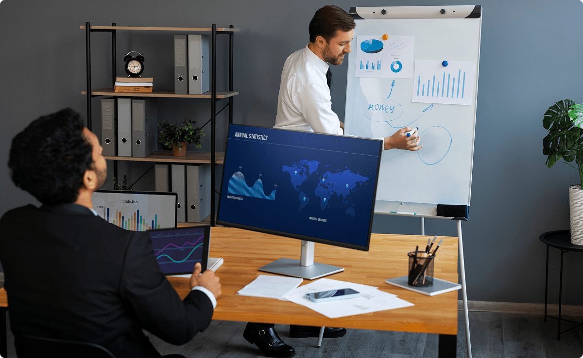 Two professionals analyzing charts and graphs on a screen and whiteboard, representing an asset management system.